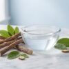 Licorice root sticks and green leaves beside a glass bowl of clear water on a marble surface